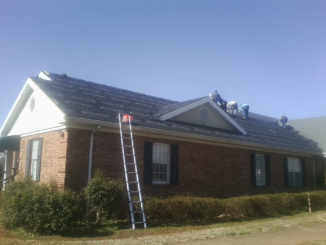 Crew tearing shingles off a brick ranch roof with a ladder set — Metal Roof Repair work by our crew in Hastings